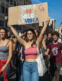 A group of young people participating in a protest or demonstration, walking down a street in a city setting with buildings in the background. The central figure, a woman, is holding up a cardboard sign with colorful text in Italian. The crowd is casually dressed, and the atmosphere suggests a peaceful and energetic gathering.