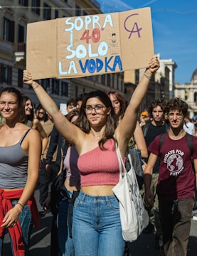 A group of young people participating in a protest or demonstration, walking down a street in a city setting with buildings in the background. The central figure, a woman, is holding up a cardboard sign with colorful text in Italian. The crowd is casually dressed, and the atmosphere suggests a peaceful and energetic gathering.