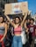A group of young people participating in a protest or demonstration, walking down a street in a city setting with buildings in the background. The central figure, a woman, is holding up a cardboard sign with colorful text in Italian. The crowd is casually dressed, and the atmosphere suggests a peaceful and energetic gathering.