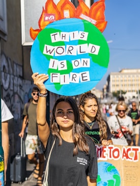 A young woman holds a colorful protest sign shaped like a globe with flames at the top, displaying the text 'THIS WORLD IS ON FIRE.' She is part of a group of people participating in a climate protest. Other participants are visible in the background, carrying signs with environmental messages. The atmosphere appears lively and engaged, with clear sunlight illuminating the scene.