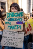 A person is holding up two protest signs made from cardboard with colorful text in Italian, expressing anti-capitalist sentiments and environmental concerns. There are other people visible in the background, and the scene appears to be a part of a larger protest or demonstration.