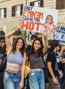 Two young women stand in the middle of a crowd holding a sign featuring a graphic image and bold text in red and blue. The background reveals a building with arched windows.
