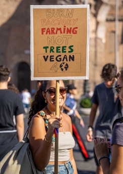 A woman is standing outside holding a protest sign. The sign reads 'BURN FACTORY FARMING NOT TREES VEGAN 4 (with an earth symbol)'. She is wearing sunglasses and smiling at the camera. There are other people in the background, indicating a public event or gathering.
