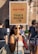A woman is standing outside holding a protest sign. The sign reads 'BURN FACTORY FARMING NOT TREES VEGAN 4 (with an earth symbol)'. She is wearing sunglasses and smiling at the camera. There are other people in the background, indicating a public event or gathering.