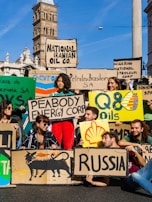 A group of people are gathered holding protest signs with names of various oil companies and countries, such as 'National Iranian Oil Co.', 'Nigerian National Petroleum Corp', 'Shell', and 'Russia'. The scene is set in an urban environment with historic architecture in the background. The individuals appear to be advocating for environmental issues related to fossil fuels.