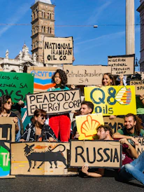 A group of people are gathered holding protest signs with names of various oil companies and countries, such as 'National Iranian Oil Co.', 'Nigerian National Petroleum Corp', 'Shell', and 'Russia'. The scene is set in an urban environment with historic architecture in the background. The individuals appear to be advocating for environmental issues related to fossil fuels.