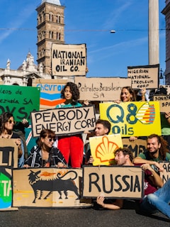 A group of people are gathered holding protest signs with names of various oil companies and countries, such as 'National Iranian Oil Co.', 'Nigerian National Petroleum Corp', 'Shell', and 'Russia'. The scene is set in an urban environment with historic architecture in the background. The individuals appear to be advocating for environmental issues related to fossil fuels.