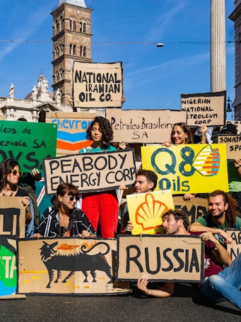 A group of people are gathered holding protest signs with names of various oil companies and countries, such as 'National Iranian Oil Co.', 'Nigerian National Petroleum Corp', 'Shell', and 'Russia'. The scene is set in an urban environment with historic architecture in the background. The individuals appear to be advocating for environmental issues related to fossil fuels.