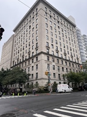 A large, rectangular, multi-story building with a classic architectural design is situated on a street corner. The facade features numerous windows and detailed stonework. Several trees and shrubs are present at the base of the building, and a few pedestrians are walking nearby. A white van labeled 'Pro-Tech' is parked alongside the street. The sky is overcast, contributing to a muted ambiance.