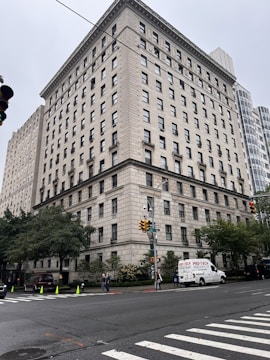 A large, rectangular, multi-story building with a classic architectural design is situated on a street corner. The facade features numerous windows and detailed stonework. Several trees and shrubs are present at the base of the building, and a few pedestrians are walking nearby. A white van labeled 'Pro-Tech' is parked alongside the street. The sky is overcast, contributing to a muted ambiance.
