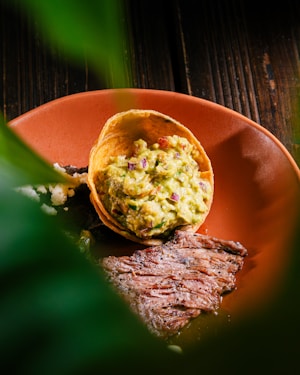 A close-up of a dish featuring a piece of grilled steak garnished with a scoop of guacamole served in a tortilla cup, set on a terracotta-colored plate. The bottom of the image includes green leaves, possibly from a plant, partially obscuring the view.