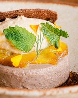 A beautifully presented dessert features a light brown mousse or cake base, topped with slices of caramelized or roasted yellow fruit, possibly pineapple. Fresh green leaves and delicate yellow flowers embellish the dish, while a textured brown wafer or cracker is placed on top.