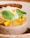 A beautifully presented dessert features a light brown mousse or cake base, topped with slices of caramelized or roasted yellow fruit, possibly pineapple. Fresh green leaves and delicate yellow flowers embellish the dish, while a textured brown wafer or cracker is placed on top.