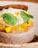 A beautifully presented dessert features a light brown mousse or cake base, topped with slices of caramelized or roasted yellow fruit, possibly pineapple. Fresh green leaves and delicate yellow flowers embellish the dish, while a textured brown wafer or cracker is placed on top.
