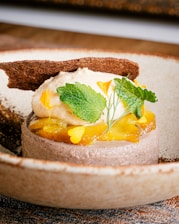 A beautifully plated dessert with a creamy base topped with citrus segments, fresh mint leaves, and delicate edible petals. A thin, crispy chocolate wafer is elegantly positioned on top, all set on a rustic ceramic plate.