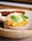A beautifully plated dessert with a creamy base topped with citrus segments, fresh mint leaves, and delicate edible petals. A thin, crispy chocolate wafer is elegantly positioned on top, all set on a rustic ceramic plate.