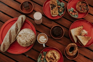 A variety of baked goods and beverages arranged on red plates on a wooden table. The dishes include a long baguette, a round loaf, slices of sandwich with coleslaw, a quiche with cherry tomatoes and greens, a pastry with filling, a slice of savory bread with toppings, a chocolate dessert, a coffee with latte art, and a creamy drink.