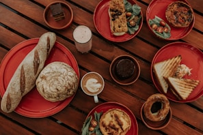 A variety of baked goods and beverages arranged on red plates on a wooden table. The dishes include a long baguette, a round loaf, slices of sandwich with coleslaw, a quiche with cherry tomatoes and greens, a pastry with filling, a slice of savory bread with toppings, a chocolate dessert, a coffee with latte art, and a creamy drink.