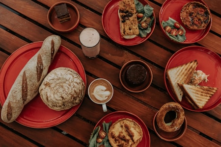A variety of baked goods and beverages arranged on red plates on a wooden table. The dishes include a long baguette, a round loaf, slices of sandwich with coleslaw, a quiche with cherry tomatoes and greens, a pastry with filling, a slice of savory bread with toppings, a chocolate dessert, a coffee with latte art, and a creamy drink.