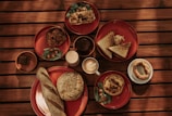 A wooden table is set with various breakfast items on red plates, including a loaf of bread, a sandwich, pastries, and a dish with greens and cherry tomatoes. There are cups of coffee and juice alongside these items.