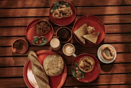 A wooden table is set with various breakfast items on red plates, including a loaf of bread, a sandwich, pastries, and a dish with greens and cherry tomatoes. There are cups of coffee and juice alongside these items.