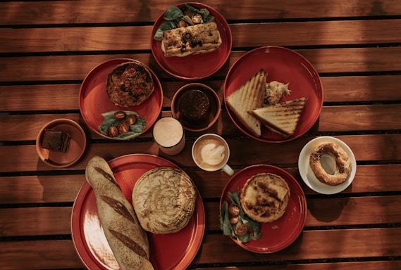 A wooden table is set with various breakfast items on red plates, including a loaf of bread, a sandwich, pastries, and a dish with greens and cherry tomatoes. There are cups of coffee and juice alongside these items.