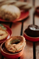 A variety of baked goods, including a round pastry with a golden crust and green filling, arranged on small, red ceramic dishes. In the background, the out-of-focus bread and a chocolate dessert add to the warm atmosphere.