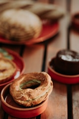 A variety of baked goods, including a round pastry with a golden crust and green filling, arranged on small, red ceramic dishes. In the background, the out-of-focus bread and a chocolate dessert add to the warm atmosphere.