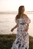 A woman in a floral dress stands on grass by a beach during sunset. Her dress features a pattern of large roses in pink and purple hues, and she appears to be enjoying the breeze as her hair flows.