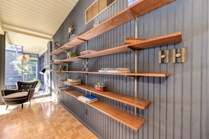 A mid-century modern interior features a dark-paneled wall with open wooden shelves mounted on it. The shelves hold various decor items such as books, plants, and small sculptures. To the left, a black modern armchair with a white cushion is placed next to large windows, which bring in natural light, creating a bright and airy atmosphere. A floor lamp stands beside the chair, and there is a parquet flooring.