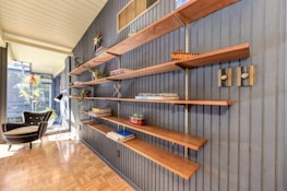 A mid-century modern interior features a dark-paneled wall with open wooden shelves mounted on it. The shelves hold various decor items such as books, plants, and small sculptures. To the left, a black modern armchair with a white cushion is placed next to large windows, which bring in natural light, creating a bright and airy atmosphere. A floor lamp stands beside the chair, and there is a parquet flooring.