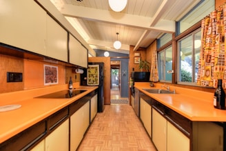 A retro-style kitchen features orange countertops, light-colored cabinets, and wood-paneled walls. The room is well-lit with a high ceiling and hanging spherical lights. Large windows on the right side are adorned with patterned curtains. A bottle of wine sits atop the counter, and there is a view of a plant and wall decorations at the far end of the kitchen.