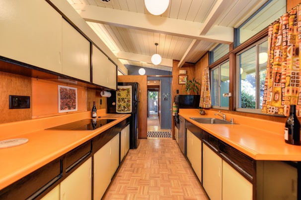 A retro-style kitchen features orange countertops, light-colored cabinets, and wood-paneled walls. The room is well-lit with a high ceiling and hanging spherical lights. Large windows on the right side are adorned with patterned curtains. A bottle of wine sits atop the counter, and there is a view of a plant and wall decorations at the far end of the kitchen.