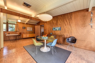 A mid-century modern dining room features wooden paneled walls and a light parquet floor. In the center, a round dining table with a white base is surrounded by colorful molded plastic chairs in hues of yellow, blue, and black. Above the table hangs a large globe-shaped pendant light. The room is furnished with a vintage sideboard and various pieces of art on the walls. A large window to the left allows natural light to enter, adorned with patterned curtains. An additional seating area with a black armchair is arranged against the wall on the right.