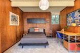 A cozy bedroom with mid-century modern style, featuring wooden paneled walls and a light gray carpet. There's a bed with assorted patterned pillows, a wall-mounted cabinet, and two colorful framed posters. A window to the right of the bed lets in natural light, and a desk with a blue chair and a yellow lamp is on the right side of the room.