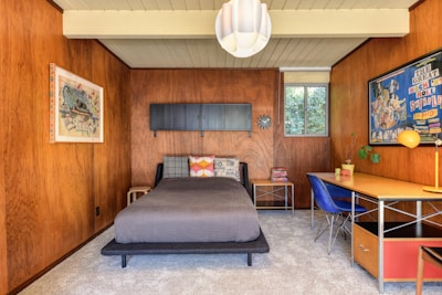 A cozy bedroom with mid-century modern style, featuring wooden paneled walls and a light gray carpet. There's a bed with assorted patterned pillows, a wall-mounted cabinet, and two colorful framed posters. A window to the right of the bed lets in natural light, and a desk with a blue chair and a yellow lamp is on the right side of the room.