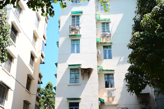 Tall residential buildings with multiple windows and green awnings are surrounded by lush green trees. The daylight creates a bright, shadowed contrast on the white walls of the buildings. Air conditioning units are visible on some of the windows.