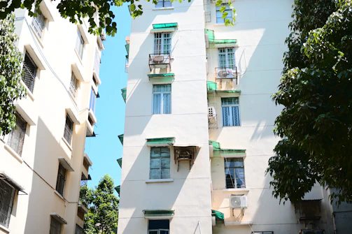Tall residential buildings with multiple windows and green awnings are surrounded by lush green trees. The daylight creates a bright, shadowed contrast on the white walls of the buildings. Air conditioning units are visible on some of the windows.