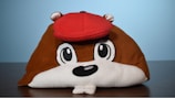 A plush toy hat resembling a cartoon animal face with large embroidered eyes and a black nose. The main body is brown with a prominent red cap on top, white underbelly, and small brown ears with white insides. The background is a simple gradient of light blue, and the hat is placed on a wooden surface.