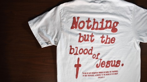 A white T-shirt with red text that reads 'Nothing but the blood of Jesus.' Below the main text, there is a smaller red biblical quote from Ephesians 1:7. The T-shirt is laid out flat on a dark wooden surface.