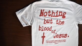 A white T-shirt with red text that reads 'Nothing but the blood of Jesus.' Below the main text, there is a smaller red biblical quote from Ephesians 1:7. The T-shirt is laid out flat on a dark wooden surface.
