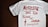 A white T-shirt with red text that reads 'Nothing but the blood of Jesus.' Below the main text, there is a smaller red biblical quote from Ephesians 1:7. The T-shirt is laid out flat on a dark wooden surface.