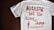 A white T-shirt with red text that reads 'Nothing but the blood of Jesus.' Below the main text, there is a smaller red biblical quote from Ephesians 1:7. The T-shirt is laid out flat on a dark wooden surface.