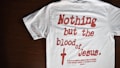 A white T-shirt with red text that reads 'Nothing but the blood of Jesus.' Below the main text, there is a smaller red biblical quote from Ephesians 1:7. The T-shirt is laid out flat on a dark wooden surface.