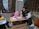A young girl wearing a pink hijab is sitting on the floor next to a man. They are both looking at a workbook on a small table. The scene appears to be in a home setting with some framed certificates and a small desk visible through a window. The setting is cozy and informal, with a rug on the floor and various study materials scattered around.
