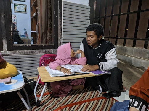 A young girl wearing a pink hijab is sitting on the floor next to a man. They are both looking at a workbook on a small table. The scene appears to be in a home setting with some framed certificates and a small desk visible through a window. The setting is cozy and informal, with a rug on the floor and various study materials scattered around.