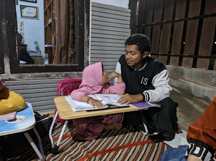 A young girl wearing a pink hijab is sitting on the floor next to a man. They are both looking at a workbook on a small table. The scene appears to be in a home setting with some framed certificates and a small desk visible through a window. The setting is cozy and informal, with a rug on the floor and various study materials scattered around.