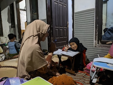 A group of people are sitting around small tables in a room. A woman and a child are prominently in the foreground. The woman is wearing a beige headscarf and appears to be interacting with the child, who is wearing a black headscarf, and smiling while holding a pencil above papers on the table. In the background, other individuals, including children, are also seated, seemingly engaged in similar activities.