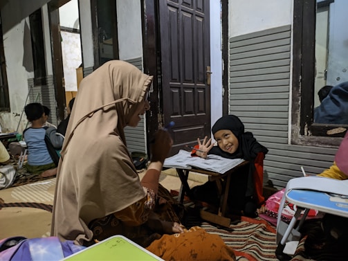 A group of people are sitting around small tables in a room. A woman and a child are prominently in the foreground. The woman is wearing a beige headscarf and appears to be interacting with the child, who is wearing a black headscarf, and smiling while holding a pencil above papers on the table. In the background, other individuals, including children, are also seated, seemingly engaged in similar activities.