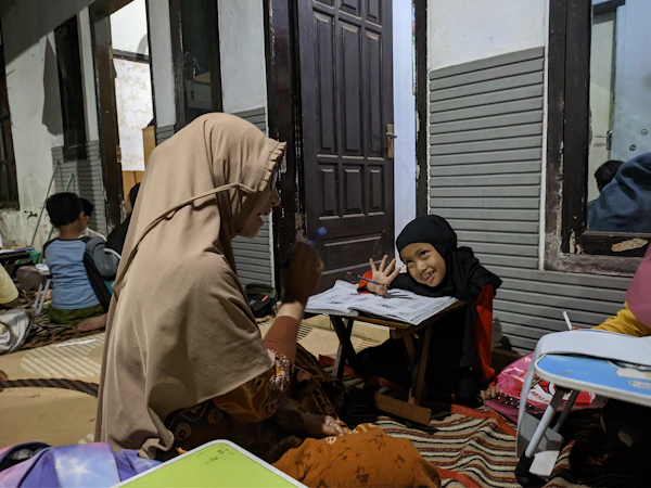 A woman in a beige headscarf helping a child write at a table during a learning session.